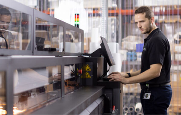 A man focused on his computer while working in a factory setting, surrounded by machinery and equipment.