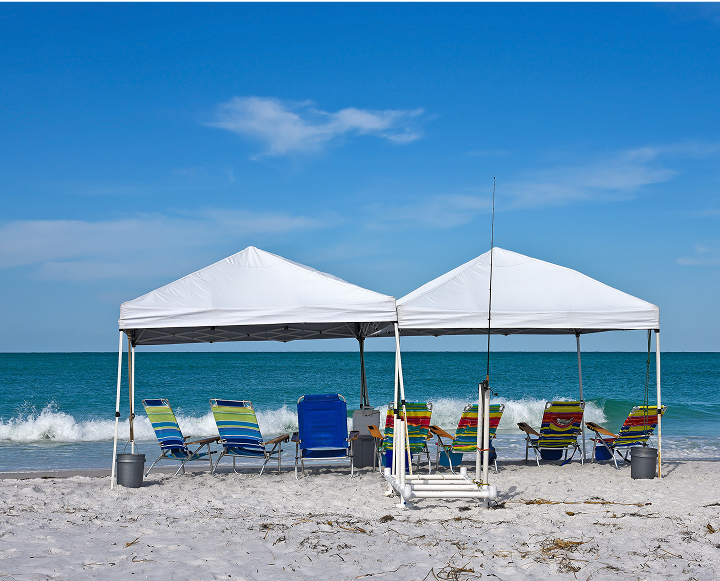 A beach scene featuring chairs and umbrellas set up under a large white tent, with the ocean in the background.