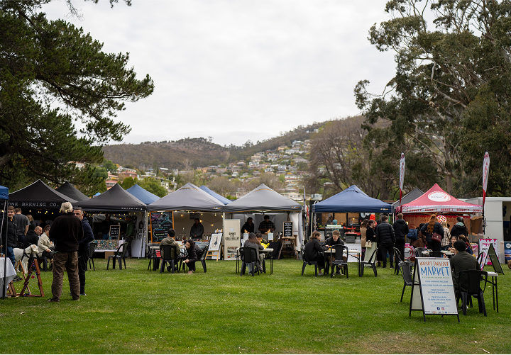 People seated at tables in a bustling outdoor market, surrounded by various stalls and vibrant displays of goods.