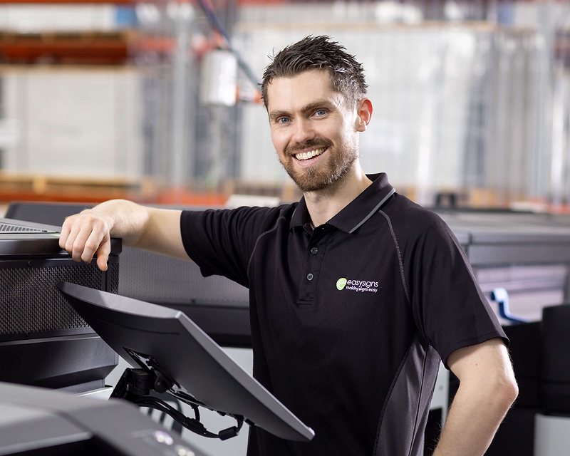 A man stands beside a large printer, observing its operation in a well-lit office environment.