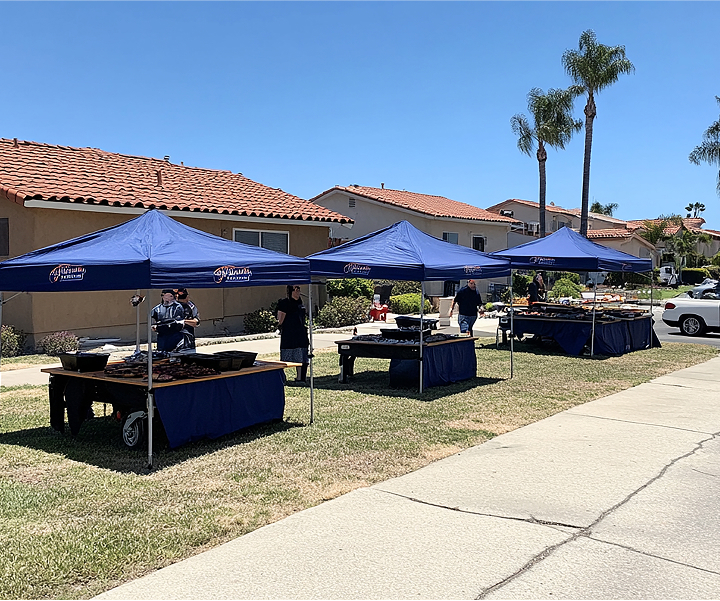 People dining at tables beneath blue tents, enjoying a meal in an outdoor setting.