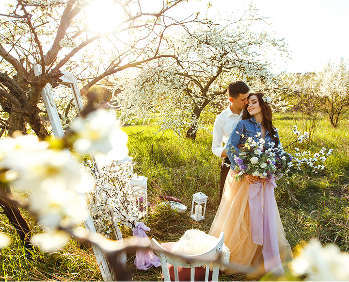 A couple stands together in a blooming orchard, surrounded by vibrant flowers and lush greenery.