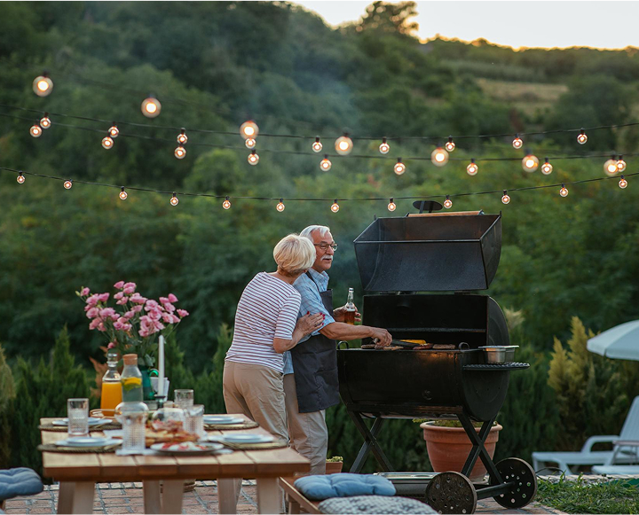An older couple enjoys grilling on a patio adorned with string lights, creating a warm and inviting atmosphere.