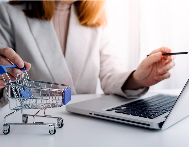 A woman sits at a table using a laptop while a shopping cart is positioned beside her.